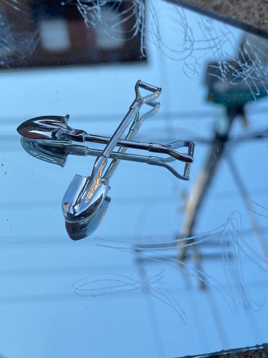 Mini sterling silver shovels are laid down in an X shape on a mirror, on a sidewalk.