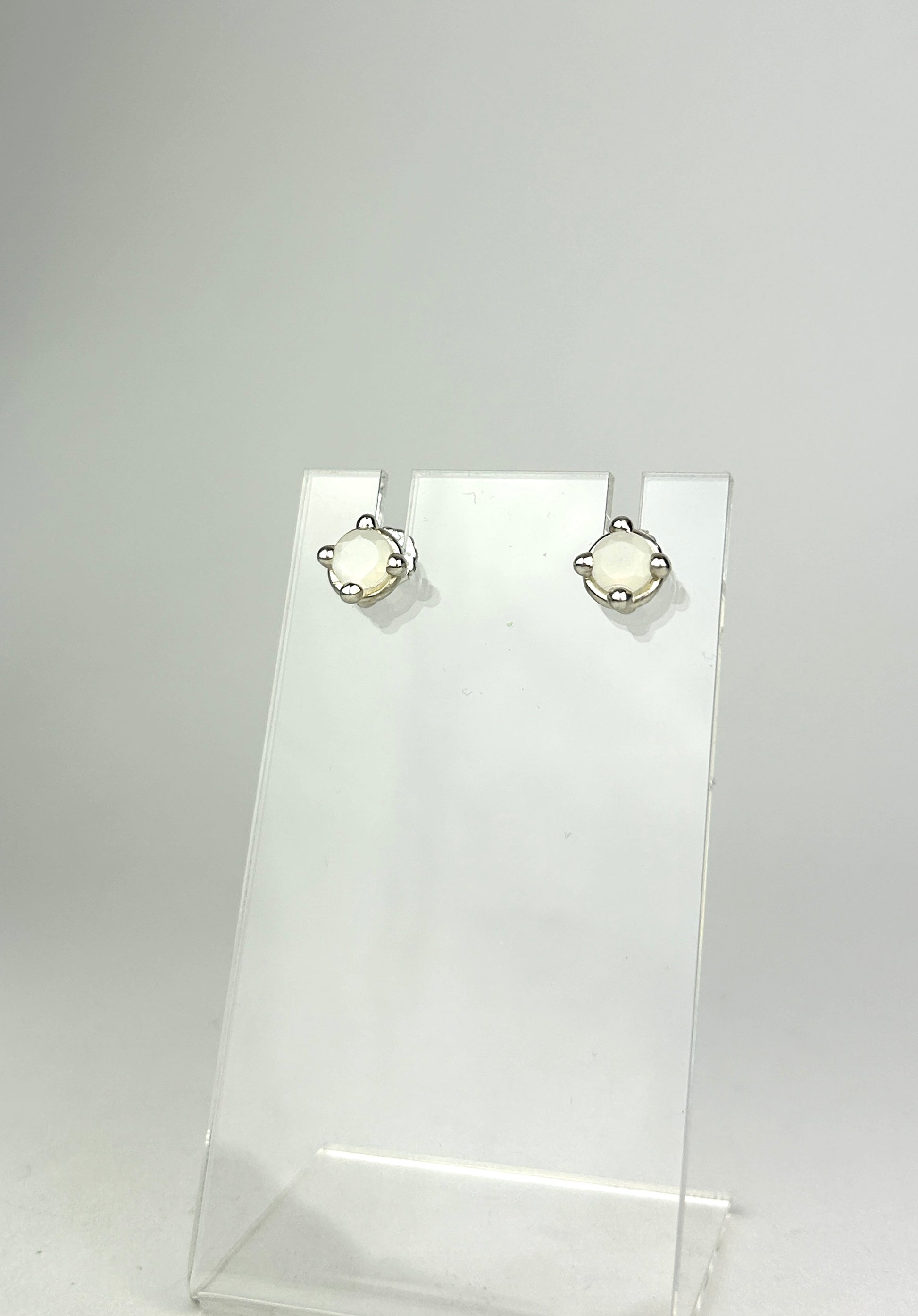 Front view of sterling silver earrings with balls for prongs, holding 6mm milky white moonstones. They are displayed on a clear acrylic jewelry display infront of a white background.