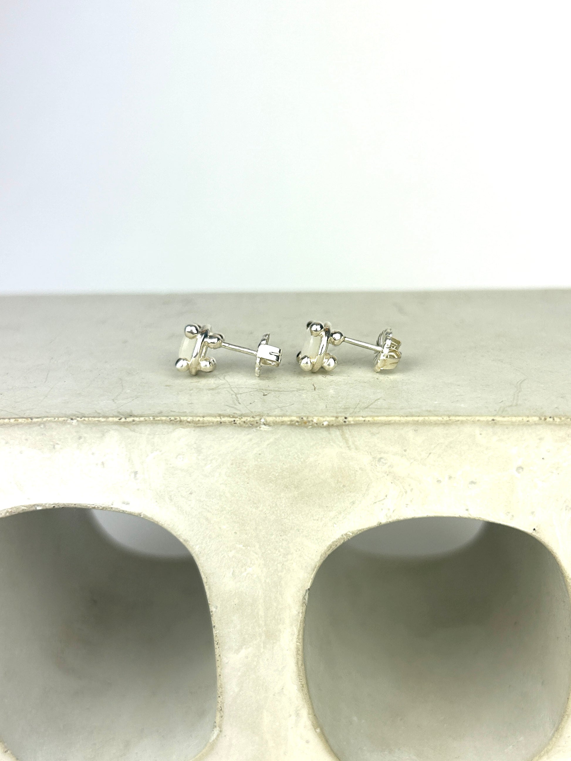 Side view of sterling silver earrings with balls for prongs, holding 6mm milky white moonstones. They are displayed laid on a small, smooth decotarive cinderblock infront of a white background.