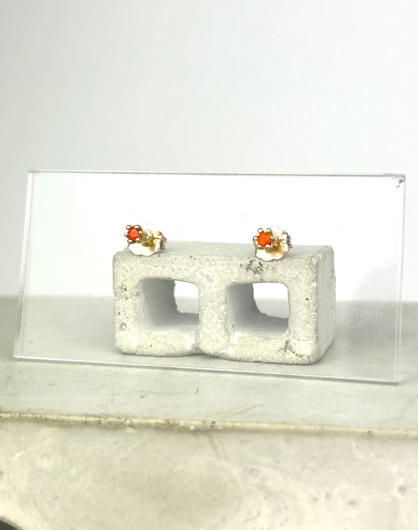 Front view 14k gold earrings with bright orange carnelians that resemble tulips are displayed on a clear acrylic card held up by a miniature cinderblock infront of a white background.
