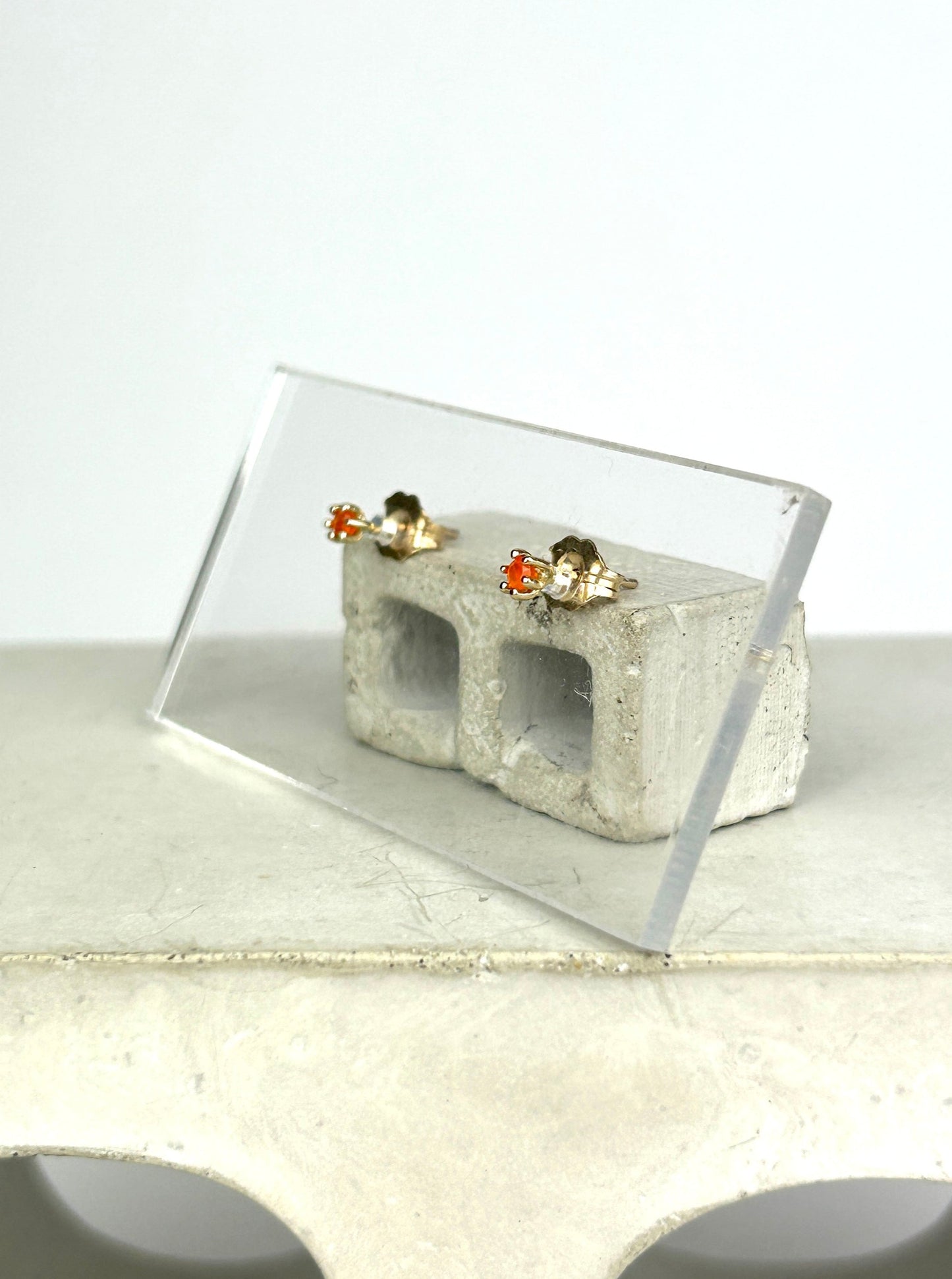 Side view 14k gold earrings with bright orange carnelians that resemble tulips are displayed on a clear acrylic card held up by a miniature cinderblock infront of a white background.