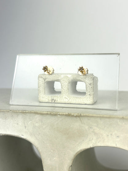 Front view 14k yellow gold stud earrings holding 2mm round cut smoky quartz displayed on a clear acrylic card learning against a miniature cinderblock on a smooth grey surface.