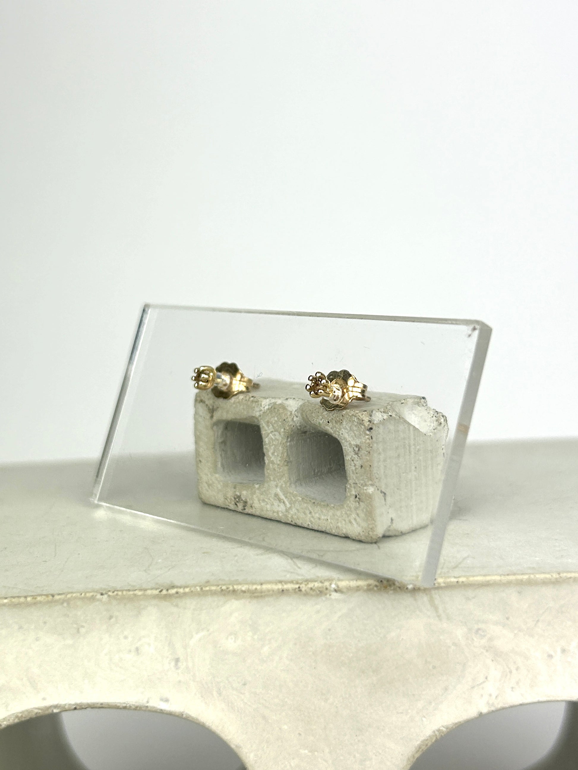 Side view 14k yellow gold stud earrings holding 2mm round cut smoky quartz displayed on a clear acrylic card learning against a miniature cinderblock on a smooth grey surface.