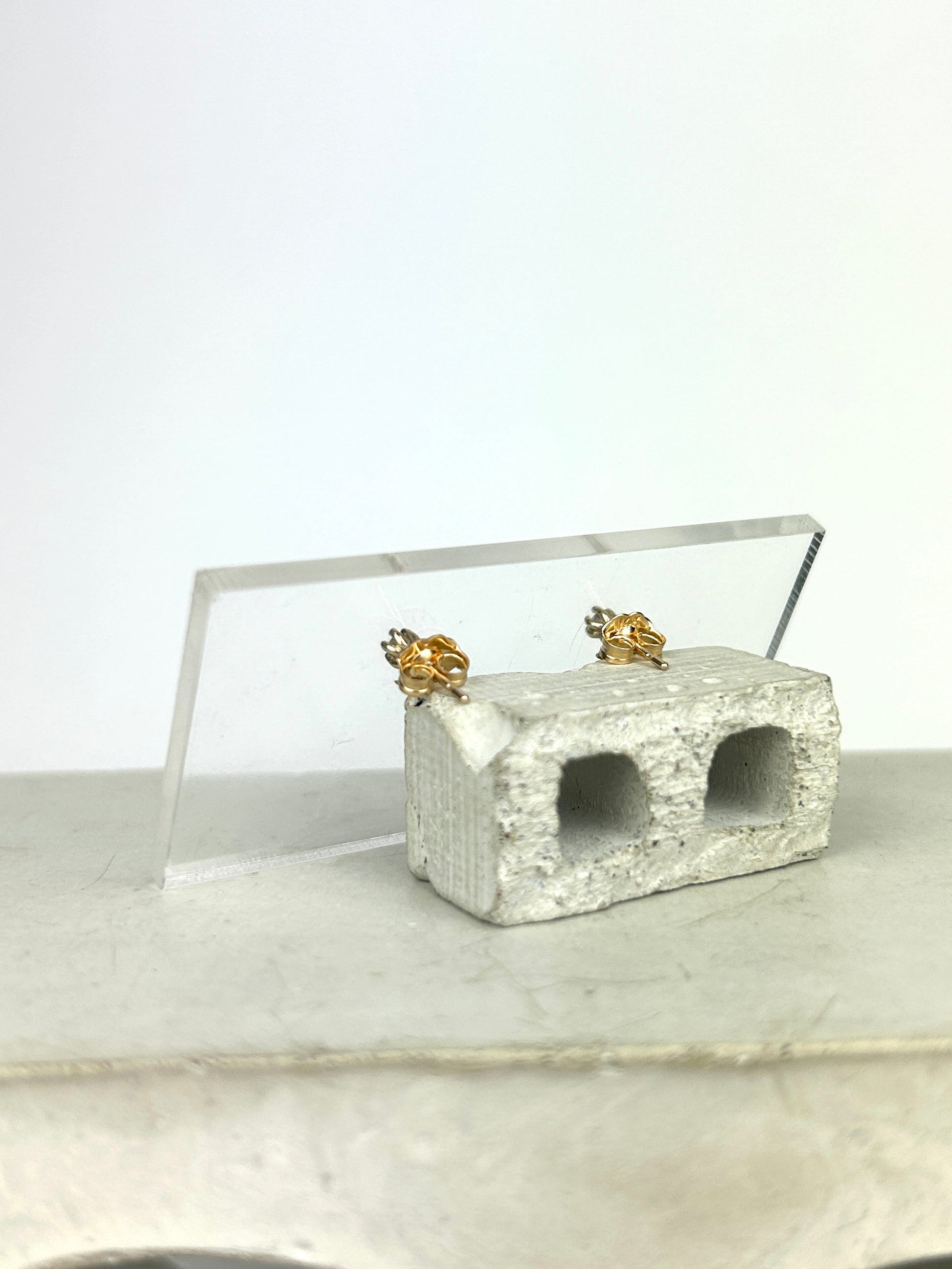 Back view 14k yellow gold stud earrings holding 2mm round cut smoky quartz displayed so you can see the earring backs on a clear acrylic card learning against a miniature cinderblock on a smooth grey surface.