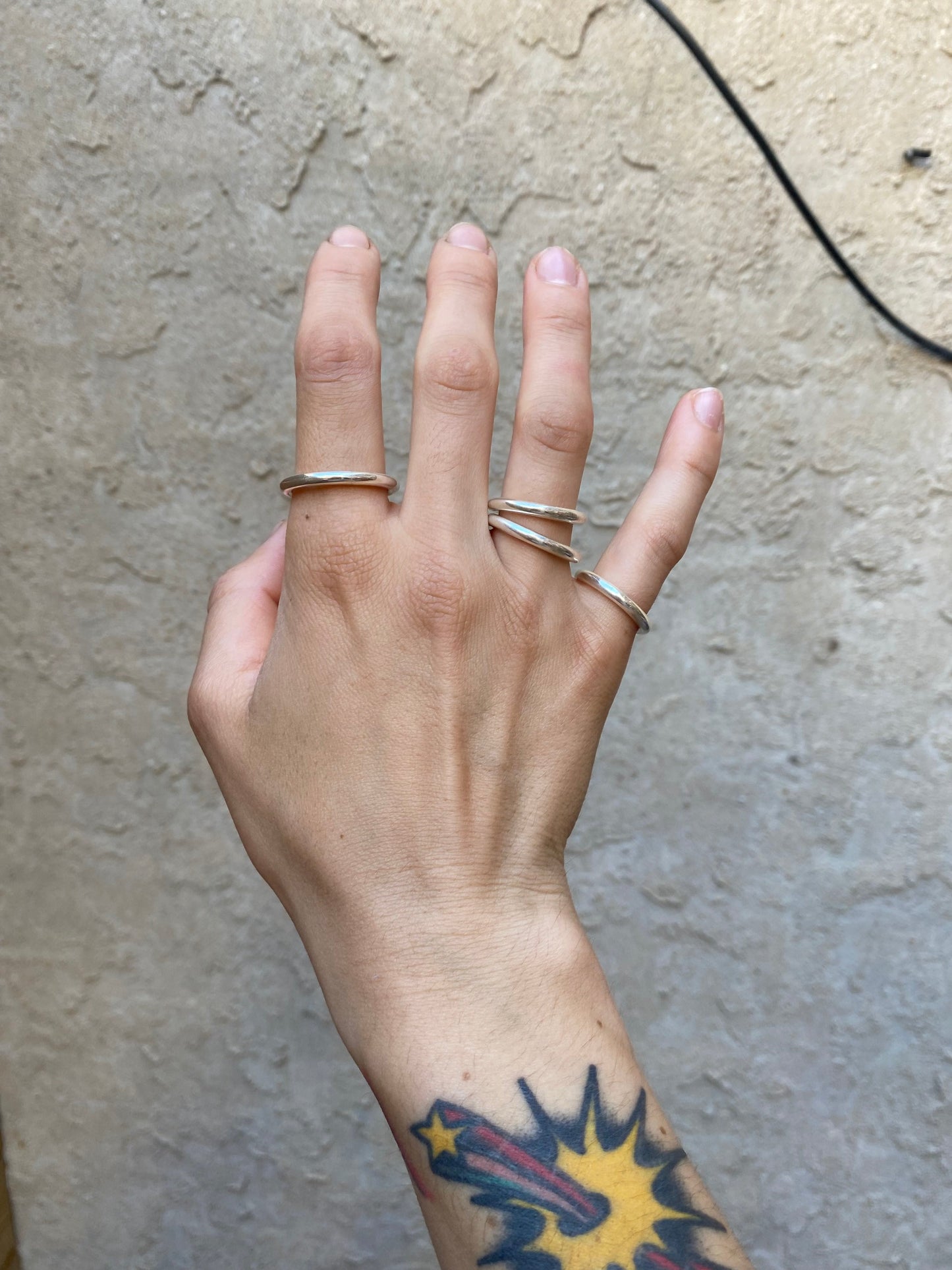 Chunky round sterling silver rings stacked on someone's right hand infront of a wall.