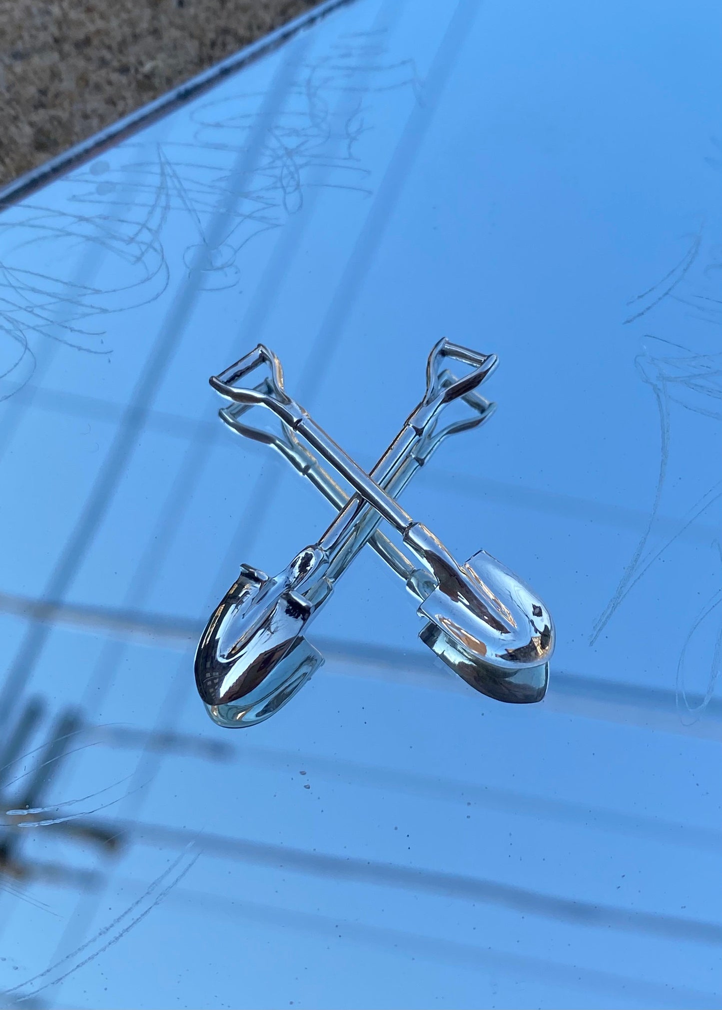 Mini sterling silver shovels are laid down in an X shape on a mirror, on a sidewalk.
